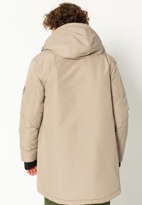 Beige gepolsterter Mantel mit Kapuze, der eine glatte Textur und einen etwas längeren Rücken aufweist, entworfen für Wärme und Komfort. Schwarze Akzente an den Bündchen.