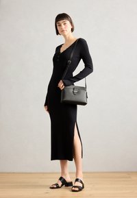 Robe noire longue à côtes avec fente latérale, présentant un décolleté en V et des détails de boutons. Assortie à un petit sac à bandoulière noir et des sandales plates.