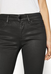 Jeans de mezclilla negra con un corte ajustado, diseño de cinco bolsillos y textura brillante. Detallados con un cierre de botón y costuras en contraste.