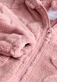 Lyserød fleecejakke med en blød, tekstureret overflade, fuld lynlås og mønstrede detaljer. Materialet ser fluffy og hyggeligt ud.
