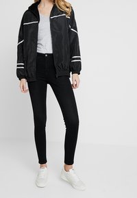 Chaqueta bomber negra con un interior texturizado y rayas reflectantes, combinada con pantalones ajustados negros y zapatillas blancas.