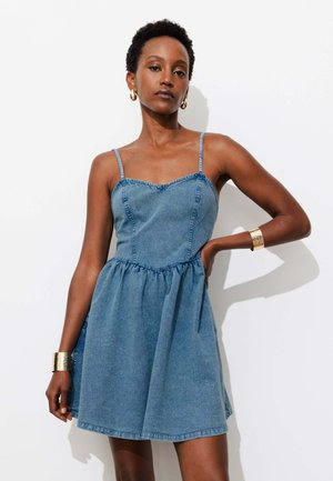 Femme portant une robe en denim bleue sans manches avec des bretelles fines, boucles d'oreilles créoles dorées et bracelets dorés, posant sur un fond blanc.