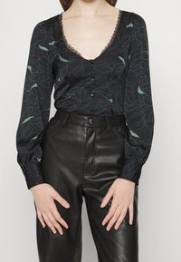 Svart blus med grönt blommönster, djup V-ringning med spetsdetaljer, långa ärmar. Matchad med högmidjade läderbyxor, slät textur.