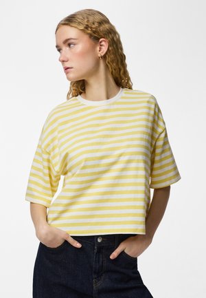 Ung kvinde med bølget blondt hår iført guldbøjleøreringe, en gul- og hvidstribet kortærmet skjorte og mørkeblå jeans med hænderne i lommerne.