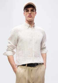 Νέος άνδρας με μανίκια γυρισμένα, φοράει μπεζ ριγέ πουκάμισο με κουμπιά, καφέ παντελόνι, μαύρη ζώνη και μπεζ καπέλο, τα χέρια στις τσέπες.