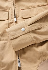 Manche de veste beige avec poignet pression, fermeture éclair avant avec tirette en métal, et poches à rabat avec détails de couture.