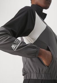 Giacca a vento grigia e nera con accenti bianchi, polsini elasticizzati, tasche con zip e logo sulla manica. Materiale morbido e leggero.