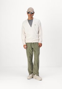 Beige strikket cardigan med V-hals, grå t-skjorte under, olivengrønne corduroybukser, beige sneakers og en beige caps med solbriller.