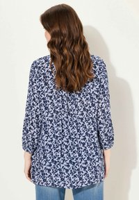 Blusa floreale blu con maniche a tre quarti, realizzata in un tessuto leggero, con una vestibilità morbida e un orlo che curva leggermente ai lati.