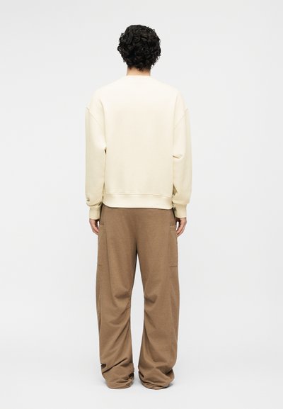 Άτομο με κατσαρά μαλλιά στέκεται με την πλάτη γυρισμένη φορώντας ένα εκρού μακρυμάνικο τοπ και χαλαρά καφέ παντελόνια σε απλό φόντο.