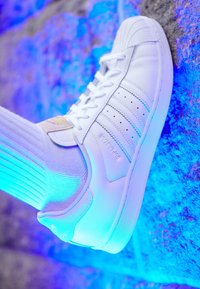 Zapatilla de cuero blanca con acentos texturizados, suela de goma contrastante y calcetín blanco acanalado. El fondo presenta una iluminación azul.