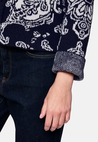 Navy patterned sweater with white designs; textured cuff with a herringbone pattern; paired with dark blue jeans, showing a hand at the cuff.