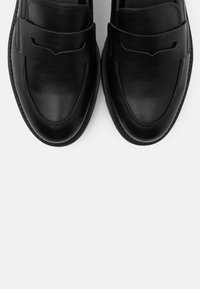 Mocassins en cuir noir avec un bout arrondi, dotés d'une bride distinctive sur le dessus et de détails de coutures subtiles le long des bords.