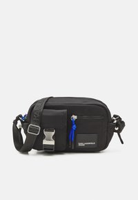 Černá nylonová crossbody taška s nastavitelným popruhem, stříbrnými kovovými prvky, přední kapsou na zip a modrými akcenty. Logo nášivka na boku. Kompaktní design.