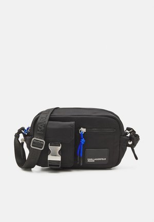 Melnas neilona plecu soma ar regulējamu siksniņu, sudraba aksesuāriem, priekšējo kabatu ar rāvējslēdzēju un ziliem akcents. Logo uzlīme sānos. Kompakts dizains.