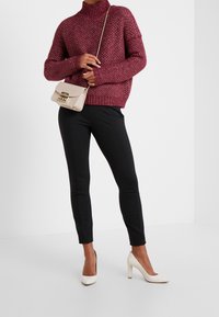 Maglione bordeaux a maglia con collo alto, pantaloni slim-fit neri e scarpe con tacco alto color crema. Piccola borsa beige con dettagli dorati.
