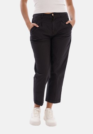 Persona che indossa pantaloni neri corti con tasche con le mani dentro, abbinati a sneakers bianche e un top bianco senza maniche.