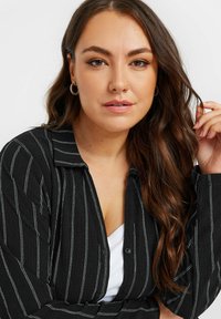 Camicia nera con bottoni e strisce verticali bianche, con collo classico e maniche lunghe. Il modello indossa un top bianco sotto.