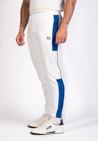Pantalons de sport blancs avec des panneaux latéraux bleus et des poignets élastiques, présentant un petit logo à l'avant et une poche zippée sur le côté.