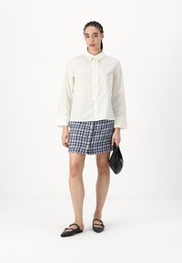 Weißes Langarmbluse mit einer Halstiefe, kombiniert mit einem gemusterten karierten Minirock und schwarzen Ballerinas, hält eine schwarze Clutch-Tasche.