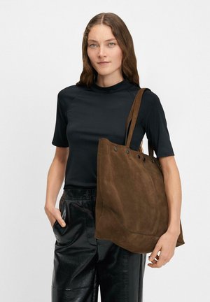 Woman wearing a black short-sleeve top and shiny black pants, carrying a large brown suede tote bag on her shoulder.