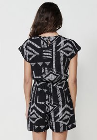 Zwarte rompert met witte geometrische patronen, korte mouwen, een striktaille en een rechte zoom. Lichte stof met textuurdetails.