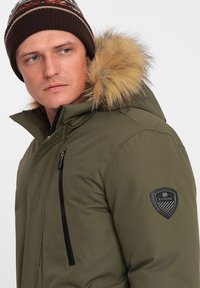 Parka verde oliva con capucha de piel sintética. Presenta cremalleras negras y un parche con el logo en la manga. La textura es suave y aislante.