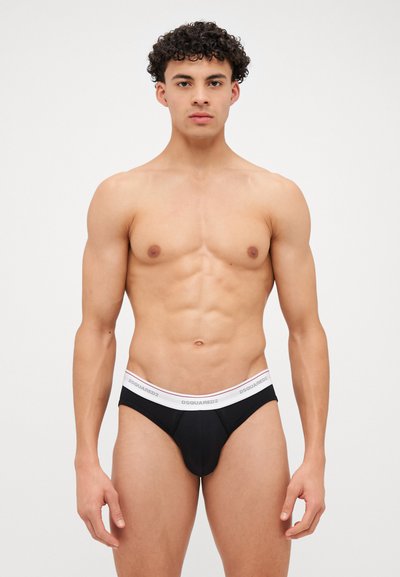 Schwarze Herren-Tanga-Unterwäsche mit einem weißen elastischen Bund, auf dem das "DSQUARED2"-Logo prangt. Glatter Stoff, körpernaher Schnitt, minimalistischer Stil.