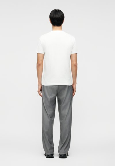 Άνδρας με μαύρα μαλλιά, γυρισμένος με την πλάτη, φοράει απλό λευκό κοντομάνικο πουκάμισο, γκρι φαρδιά παντελόνια και μαύρα παπούτσια σε λευκό φόντο.