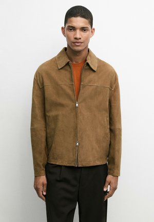 Man staat met het gezicht naar voren, draagt een bruine suède jas met ritssluiting over een oranje trui en zwarte broek tegen een effen witte achtergrond.