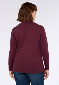 Burgundy stickad tröja med en ribbad krage och avslut, långa ärmar och en något förlängd baksida, i kombination med blå denimjeans.