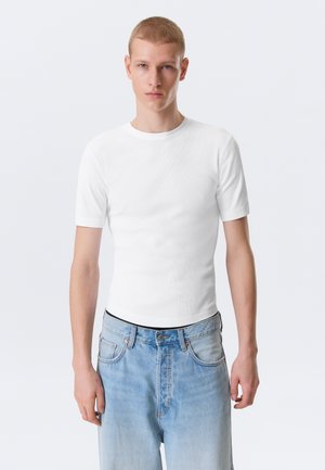 Giovane uomo con capelli rossi corti che indossa una maglietta bianca a maniche corte semplice e jeans azzurri, in piedi contro uno sfondo bianco.