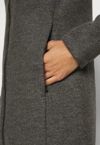Cappotto di lana grigio con una trama liscia, caratterizzato da una tasca laterale e dettagli di cucitura sottili lungo le cuciture. Chiusura con zip visibile.