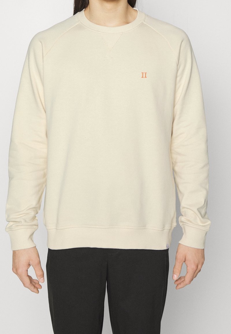 Personne portant un sweat-shirt crème à manches longues avec un petit "II" orange sur la poitrine gauche, assorti à un pantalon noir.