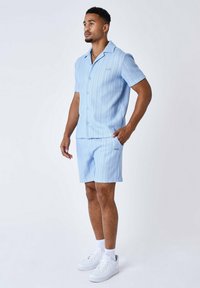 Camicia azzurra a maniche corte con bottoni e righe verticali, abbinata a pantaloni corti coordinati. Indossata con scarpe da ginnastica bianche e un design texture.