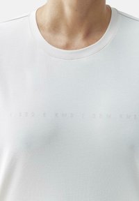 Balta medvilnė marškinėliai su apvaliu kaklu, priekyje su iškiliais užrašais: "1.332 B KM3 | 35M KM3" ir lygiu, tekstūruotu audiniu.