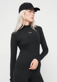 Ženska z dolgimi blond lasmi nosi črno kapo, oprijet črni dolgi rokav s tekstom "LeGer" in črne legice, gleda na stran.