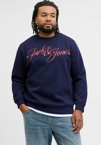 Sudadera azul marino con el logotipo "Jack & Jones" en script rojo, con puños y dobladillo acanalados, combinada con jeans de color azul claro.