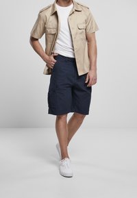 Chemise beige à manches courtes avec épaulettes, t-shirt blanc en dessous, shorts cargo navy avec poches, et baskets blanches. Design minimaliste.