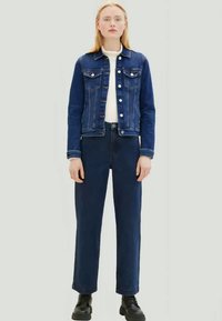 Donna in piedi con una giacca di jeans blu, jeans blu scuro, top bianco e stivali neri su uno sfondo chiaro uniforme.