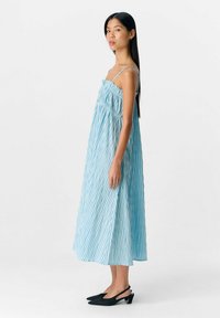 Robe maxi rayée bleu clair et blanche avec des bretelles fines et un corsage froncé. Portée avec des chaussures à talon noir. Texture lisse.