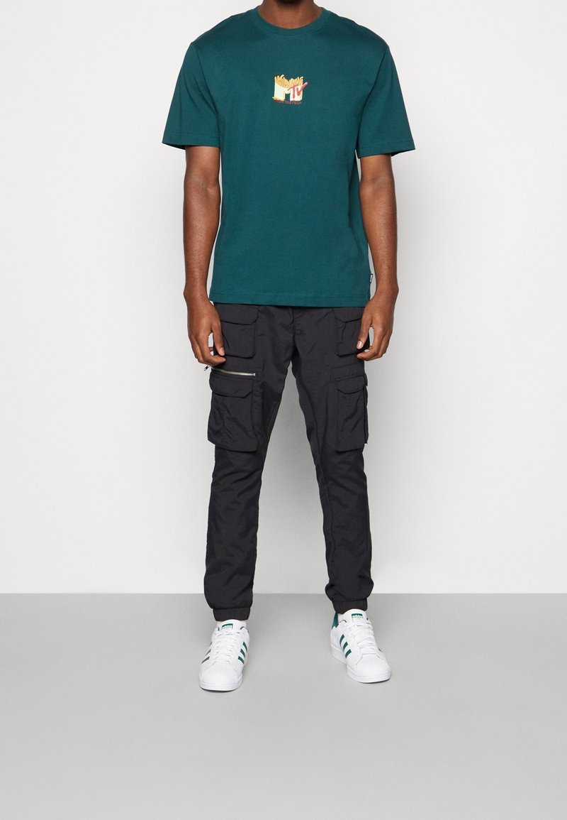 Camiseta de cuello redondo color azul verdoso con un gráfico de papas fritas y el logo de MTV, combinada con pantalones cargo negros y zapatillas blancas con acentos verdes.