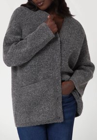 Cardigan gris foncé en laine douce, présentant un col en V, des boutons à l'avant et deux poches latérales. La conception a une coupe décontractée et une surface texturée.