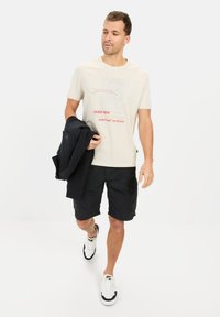 Beige grafisk T-shirt med rød tekst, sorte cargo shorts og hvide sneakers med marineblå detaljer. Modellen bærer en sort jakke.