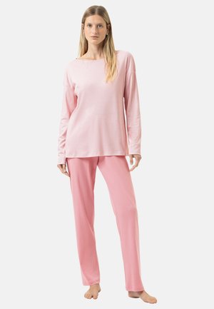 Jonge vrouw staat blootsvoets, gekleed in een lichtroze top met lange mouwen en bijpassende roze broek tegen een effen achtergrond.