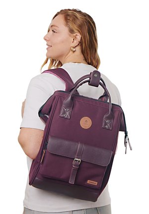 Frau mit einem lila rechteckigen Rucksack mit Lederakzenten, oberen Griffen und einer vorderen Schnallen-Tasche, im weißen Hemd nach links schauend.