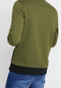 Felpa verde oliva con colletto rotondo e orlo nero, realizzata in materiale morbido. Presenta maniche lunghe e un design semplice sul retro.