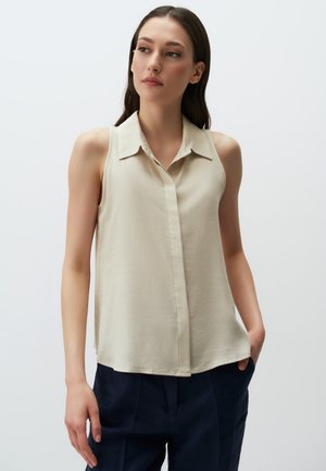 Blusa beige senza maniche con colletto e chiusura a bottoni, realizzata in tessuto morbido, abbinata a pantaloni blu navy.
