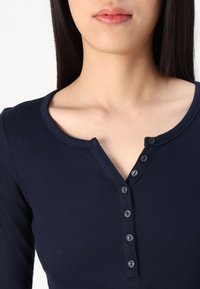 Mornarska rebrasta majica dugih rukava s okruglim izrezom i sedam crnih gumba sprijeda. Glatka tekstura materijala.
