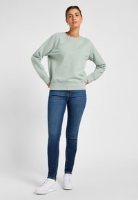 Sweatshirt vert clair avec un col rond et des poignets côtelés, associé à un jean skinny bleu et des baskets blanches. Design simple, sans motifs.
